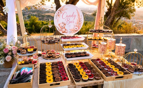 Υπαίθρια παρουσίαση candy bar με ποικιλία γλυκών σε δίσκους, ροζ πινακίδα Candy Bar, ορεινό τοπίο και διακοσμητικό στήσιμο κάτω από διάφανο υφασμάτινο κιόσκι