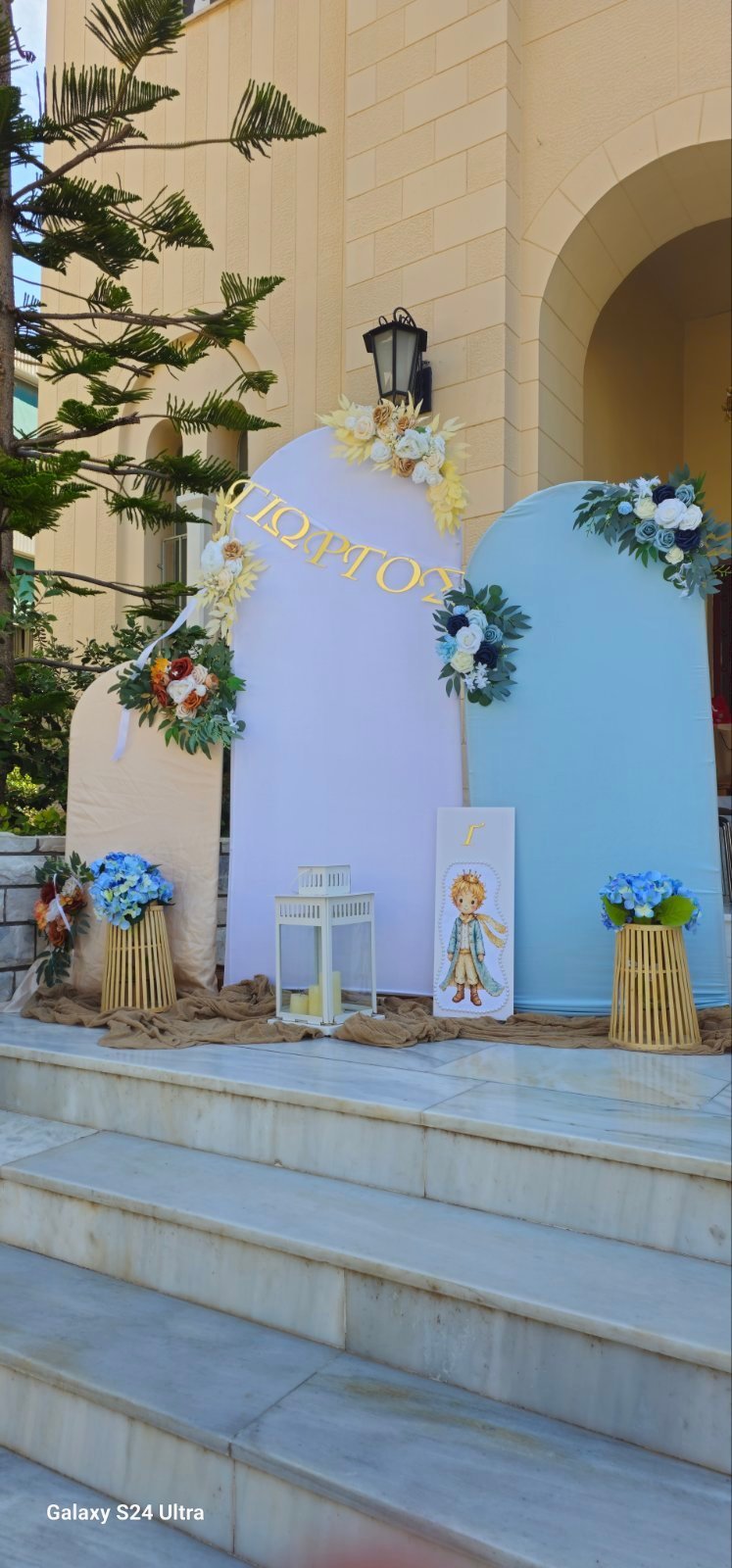 Υπαίθρια διακόσμηση με μπλε και μωβ πάνελ στολισμένα με λουλούδια, με απεικόνιση χαρακτήρα και φοίνικες μπροστά από πέτρινο κτίριο